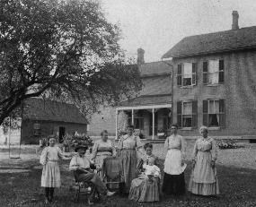 Black and white photo of the Coonrad family of eight posing in front of their farm house. The farm is a two story brick building with a covered porch and a large, grassy front yard.