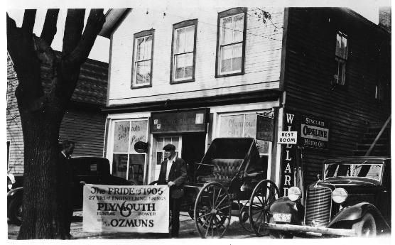 Black and white photo of two men holding a banner in front of a store with vehicles; the banner and store windows include the name "Plymouth".