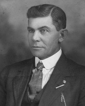 Black and white photo of Eugene Cranz, an unsmiling white man with thick, dark hair parted on the side, wearing a suit and tie with various pins on his lapel.