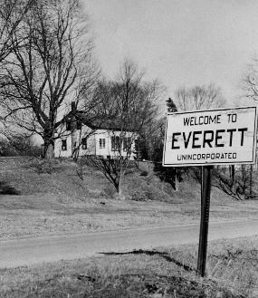Black and white photo of a white house on a hill with a sign reading "Welcome to Everett., Unincorporated" in the foreground.