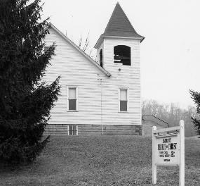Black and white photo of Everett Church of Christ, a white church with a small bell tower and steeple. A lone evergreen tree is in the foreground and a sign is in the church yard.