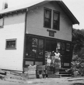 Black and white photo of Kepner's Store, Everett. A two-story house with white siding and large windows on the first floor. Wooden crates are stacked on the ground in front and on the step. A person is behind a stack of crates as tall as them.
