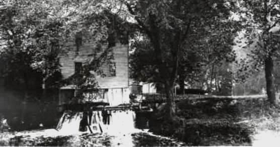 Black and white photo of a two-story white building surrounded by trees; in the foreground a waterfall spills from below the building.