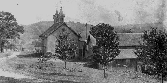 Black and white photo of a barn with two steeples; young trees line a drive in the foreground.