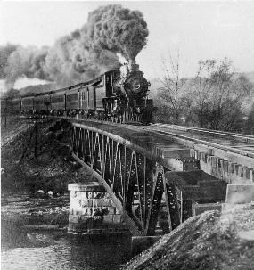 Black and white photo of a locomotive crossing a river on a narrow, wooden bridge. Dark plumes of smoke trail behind it. Some trees are in the background.