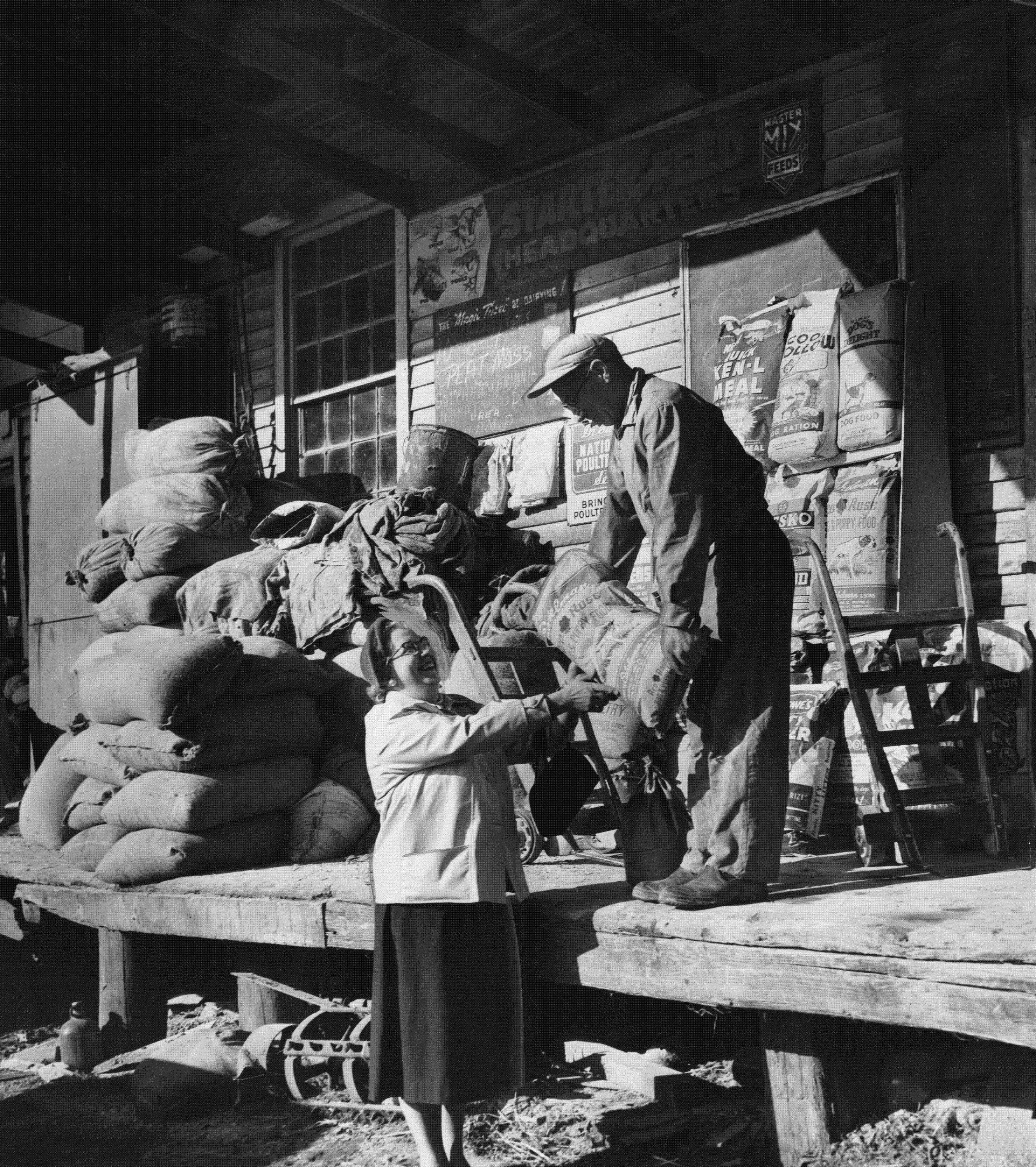 Black and white photo of a worker on a porch handing a bag of dog food to a woman standing on the ground below.