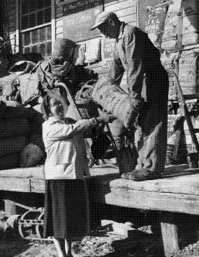 Black and white photo of a customer buying goods at Wilson's Mill, 1957. A woman is standing on the ground with a short porch at hip height and a man hands a large bag to her.