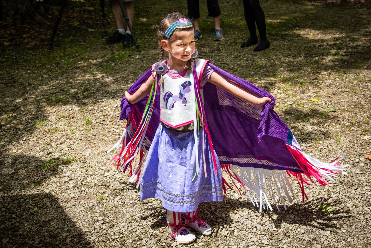 Young girl in a costume stands smiling, holding her fringed purple cape open. Unicorns decorate her white and pink overshirt and shoes. She wears a long, belted skirt, beaded headdress and round beaded ornaments with streaming ribbons in her hair.