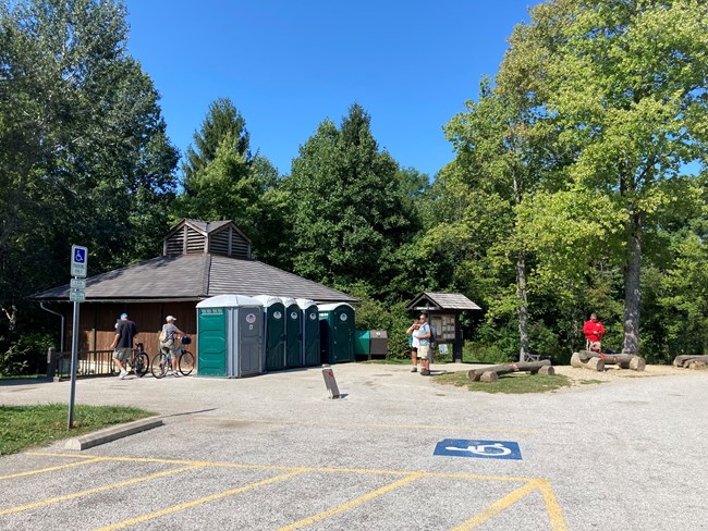 A small brown wooden building is along a paved path and parking lot with a bike rack and four portable restrooms in the front and green forest in the background.