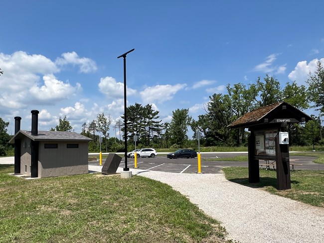 Wooden kiosk labeled "Stanford" stands along a gravel path next to a parking lot and restroom.