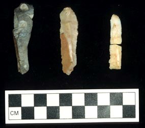 Three narrow stone tools arranged on a black background with a black and white checkered scale at the bottom.