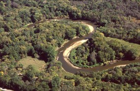 Aerial view of a brown river winding its way through green trees and shrubs; the river's course is a backwards S shape.