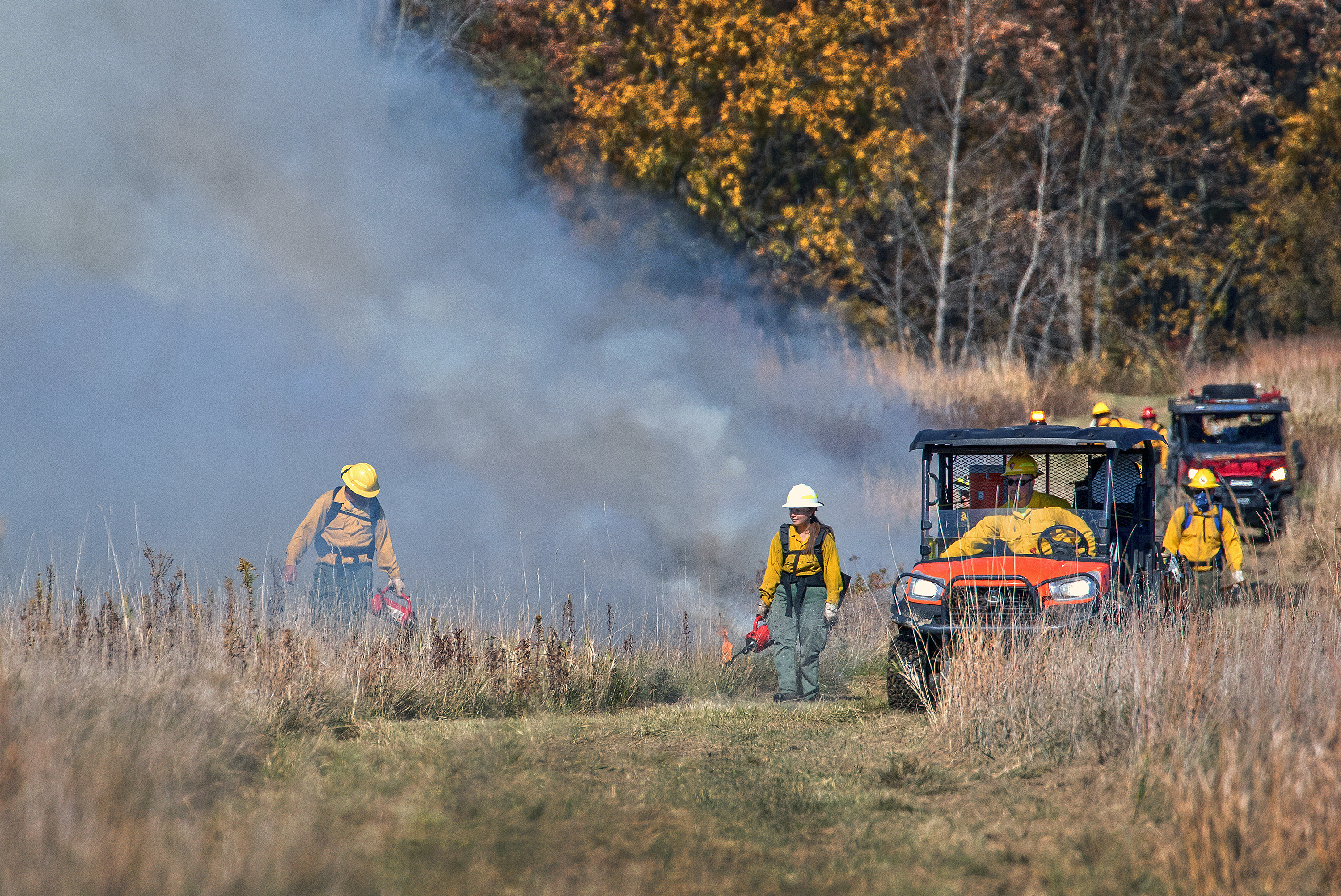 2022 Coliseum burn Several people in bright yellow shirts walk and ride in four-wheelers through a dry brown field, starting a smoldering fire, gray smoke rising off to the left.