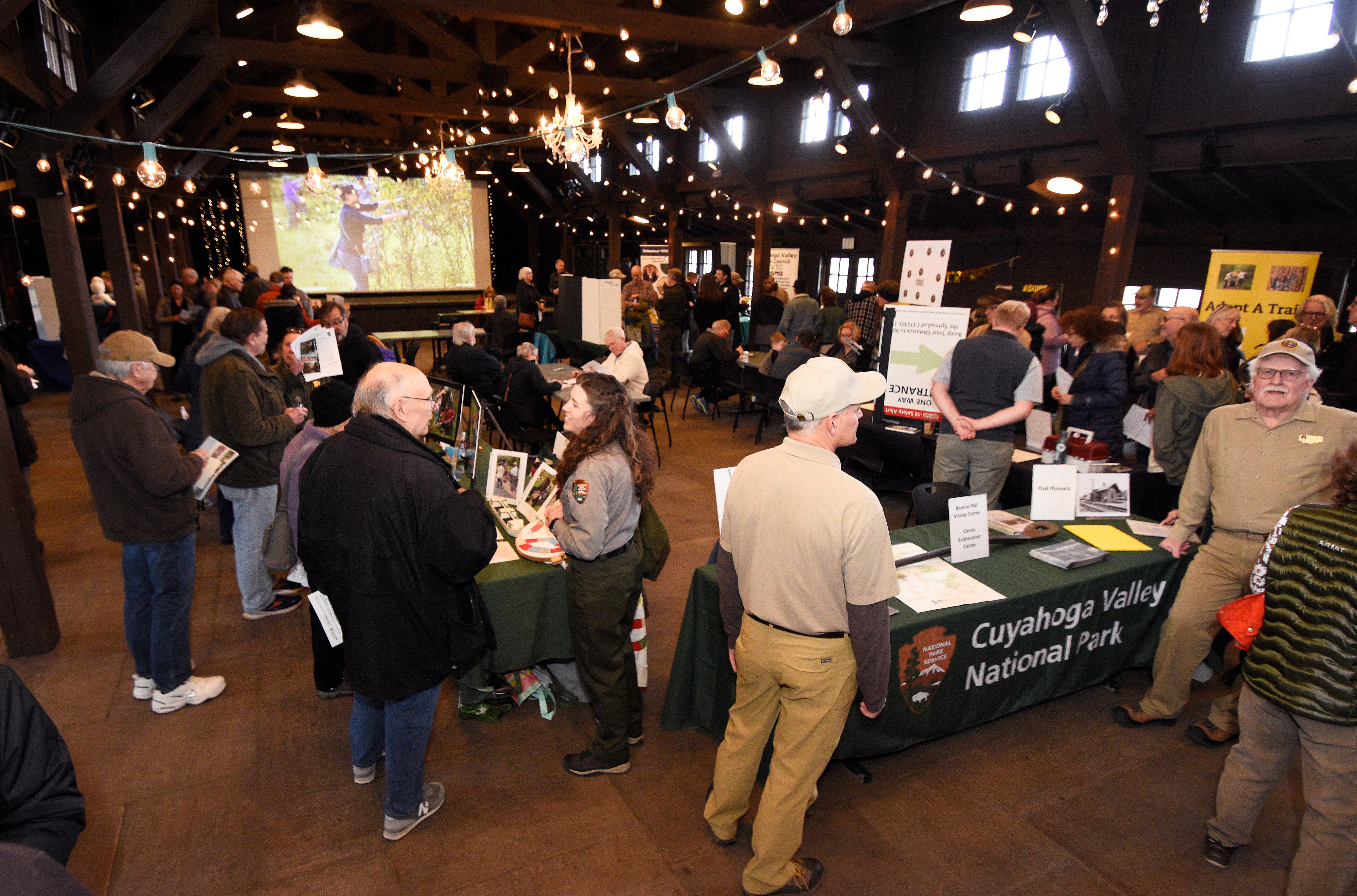 Dozens of people mingle in a large room, standing around tables, some with green table cloths that read "Cuyahoga Valley National Park."