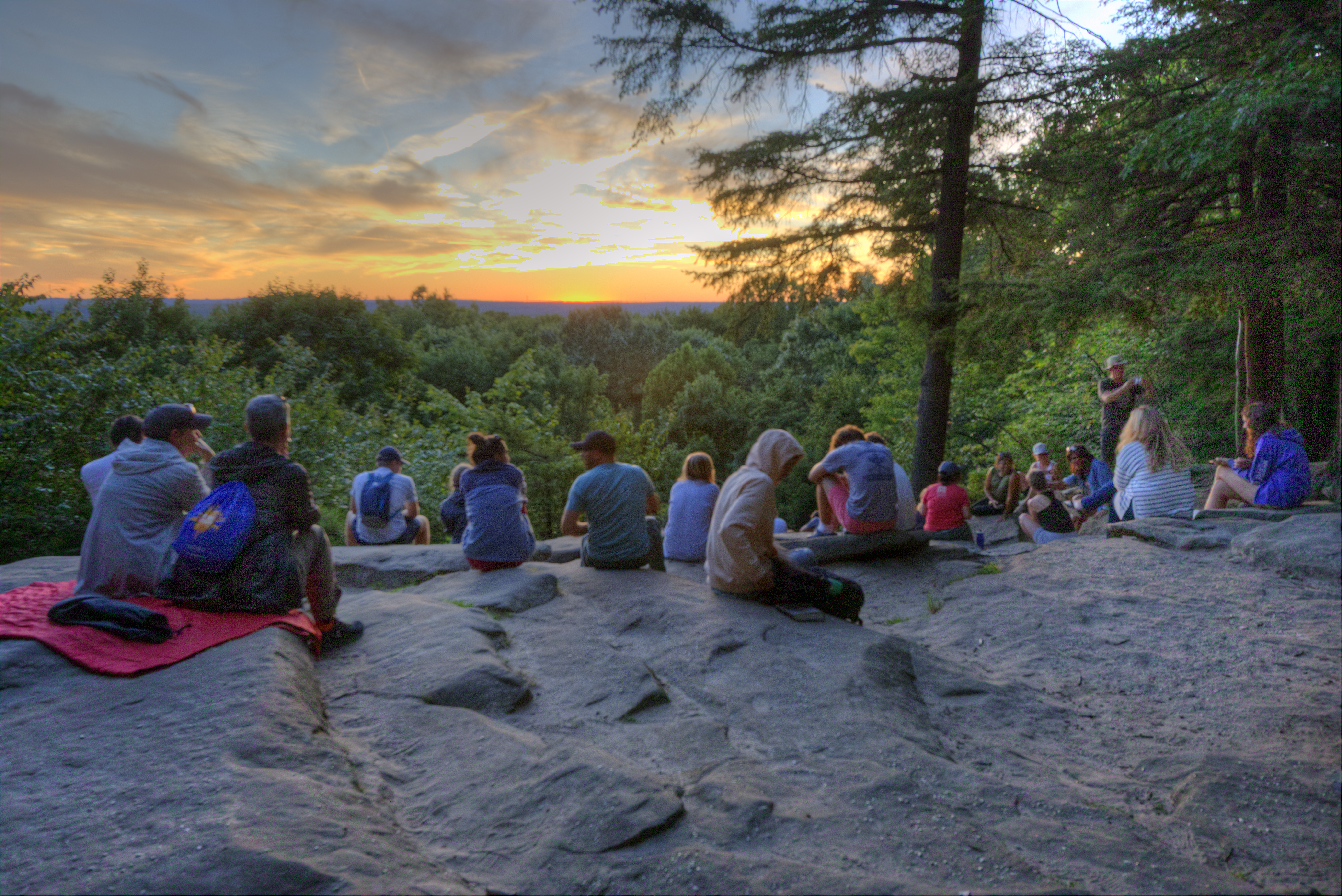 People sitting on a rock ledge at sunset overlooking a green forest