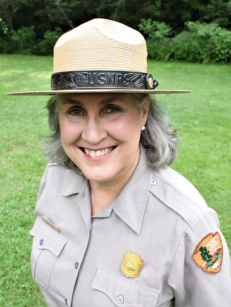 Portrait of a smiling uniformed ranger with shoulder-length dark gray hair; she wears a tan straw uniform hat and gray shirt with gold badge and National Park Service arrowhead patch on the sleeve.