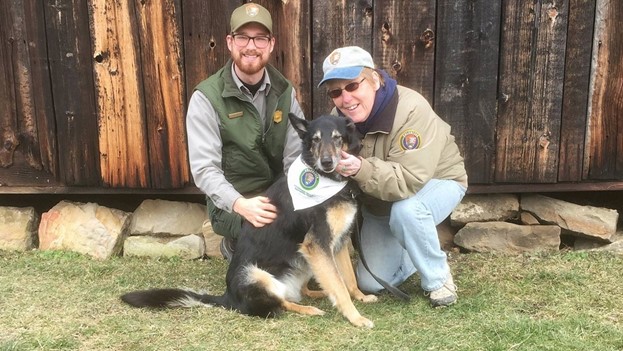 Volunteer and park ranger kneel with dog on grass in front of a wooden barn.