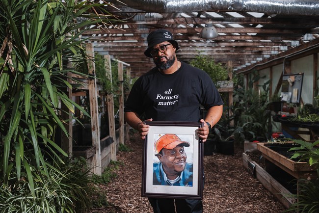 A Black man with an “all Farmers Matter” t-shirt stands in a greenhouse holding a painted portrait.