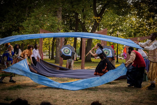A multi-racial group of young actors perform in park, undulating four large blue sheets of fabric.