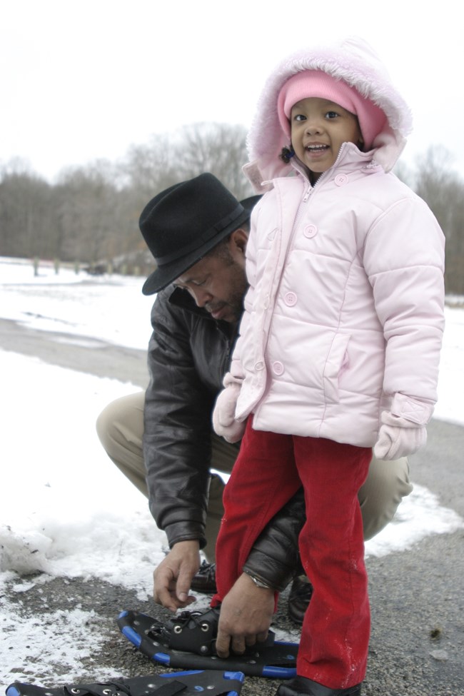 Young girl bundled in pink winter coat and red pants stands with her foot in a blue snowshoe; a man in a black hat squats to tie her shoe.