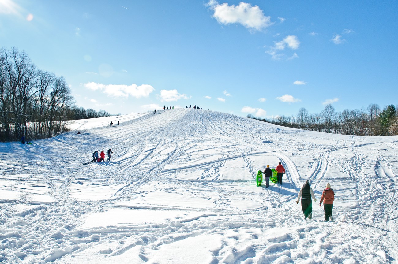 People walking up a sledding hill on a sunny snowy day.