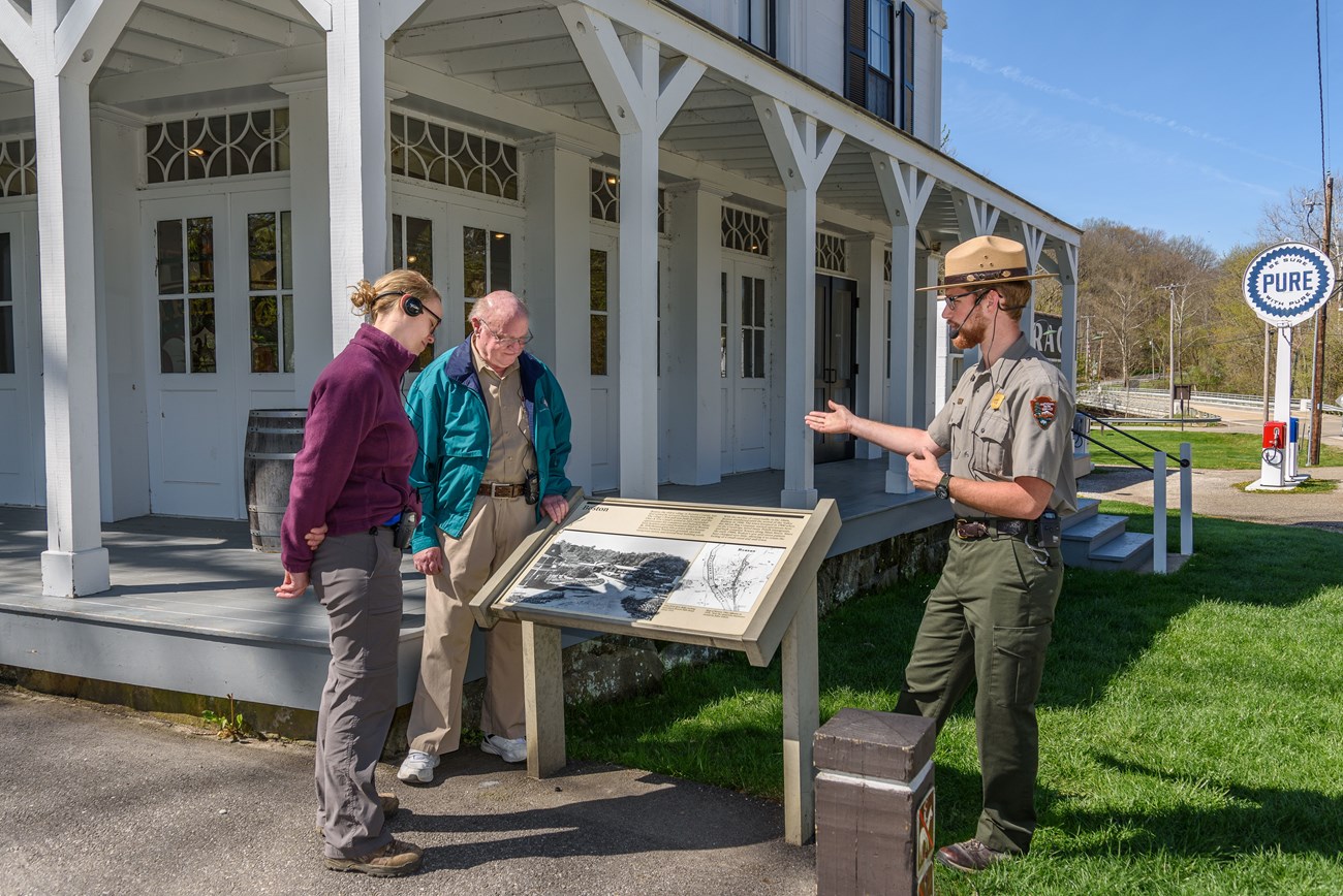 A uniformed ranger talks to two people through headsets; they stand at a panel by the porch of a white historic building with many windows.