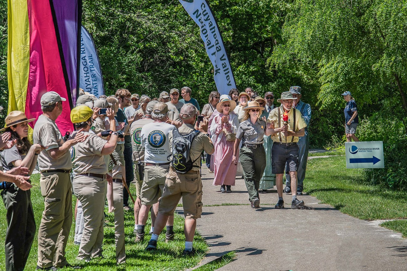 Volunteers and rangers stand and applaud as a ranger and volunteer lead a parade carrying a red-orange facsimile of a torch.