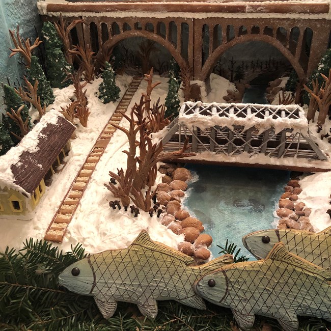 Winter scene made of gingerbread and icing. Railroad tracks and a river run under two arches of a high road bridge, shown in the background. A yellow train station is left the tracks. To the right, a smaller silver truss bridge spans the river in the midd