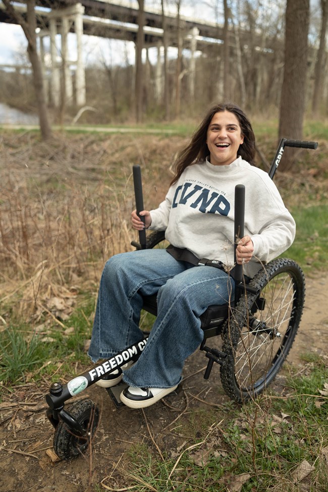 A smiling person sits in a wheelchair equipped with vertical handles and a small front third wheel, on a dirt path through trees.