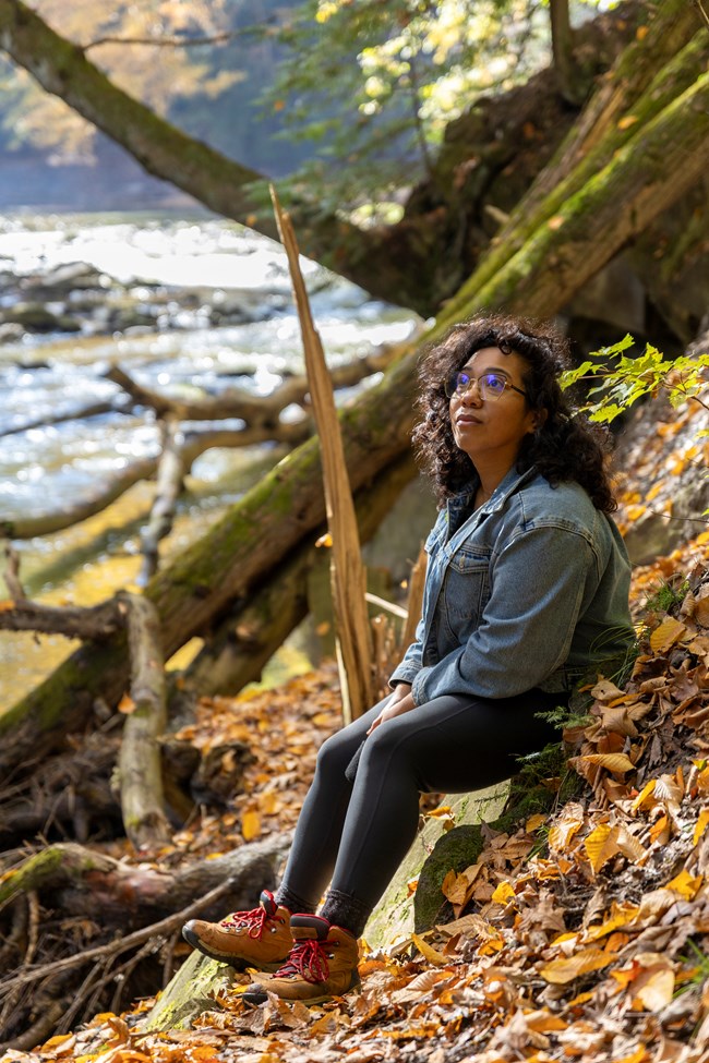 A Black woman wearing a jean jacket and hiking books sits on a riverbank, looking upward.
