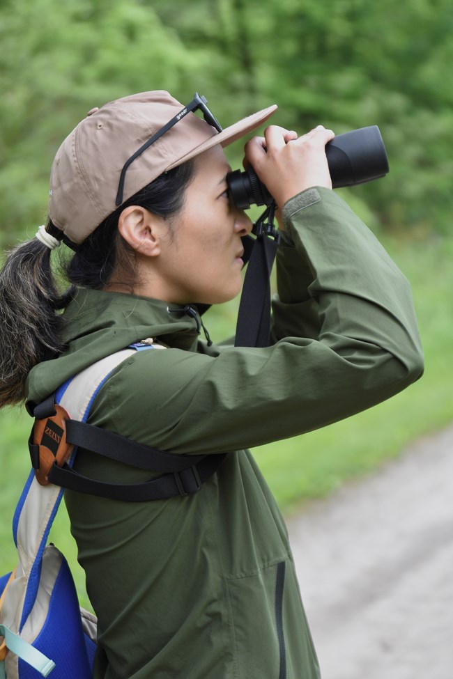 An Asian woman in a green jacket stands in profile along a trail, looking up through binoculars.
