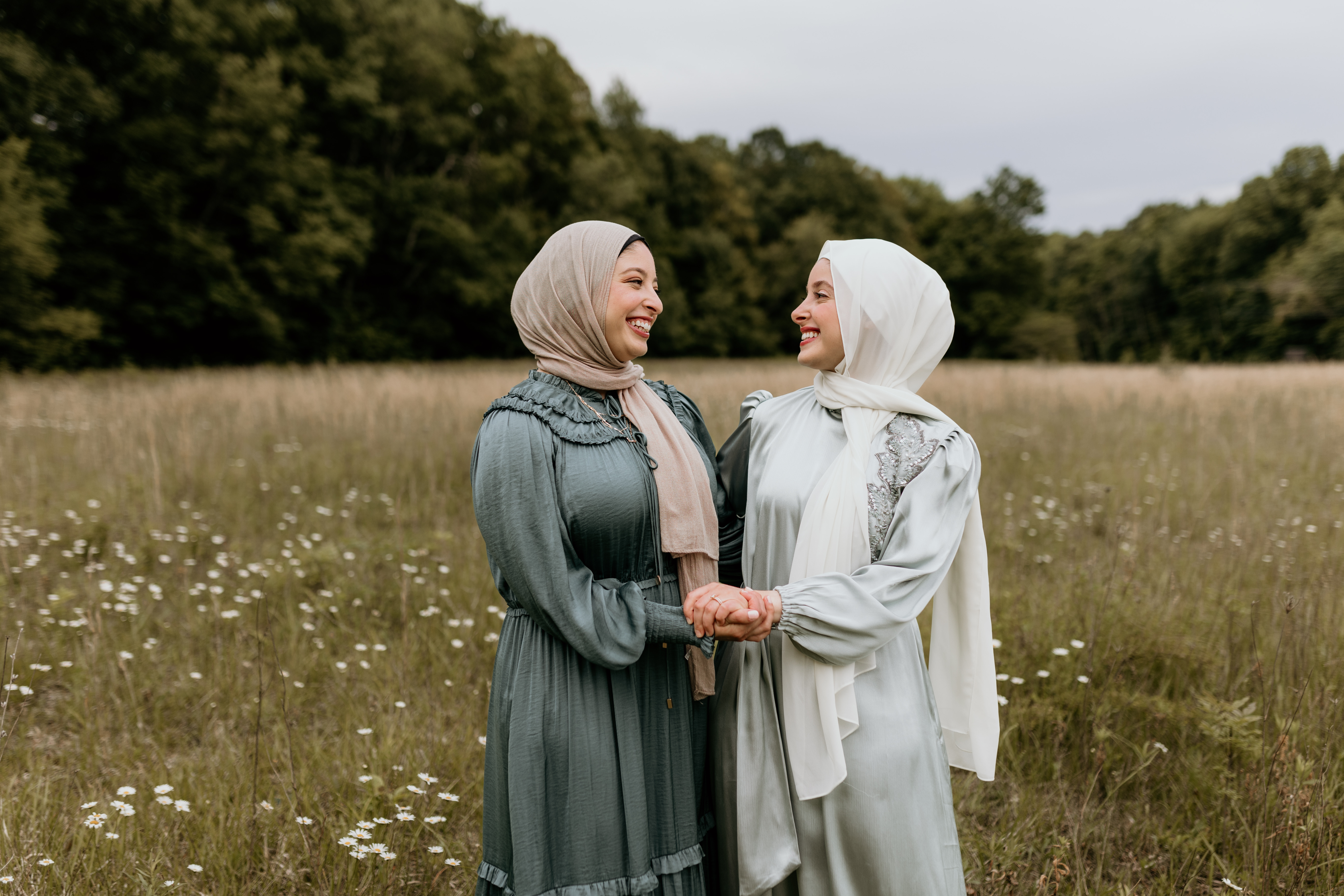 Two young women wearing head coverings and long dresses smile at each other and hold hands in a park field.