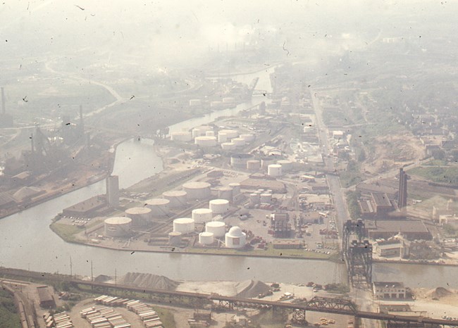 A hazy view of industry along the river: large white cylindrical tanks, railroad bridges and smokestacks dominate the landscape.