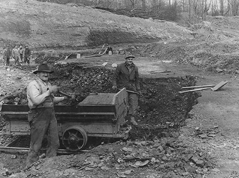 Black-and-white phot of two men shoveling coal form an open pit into a cart.