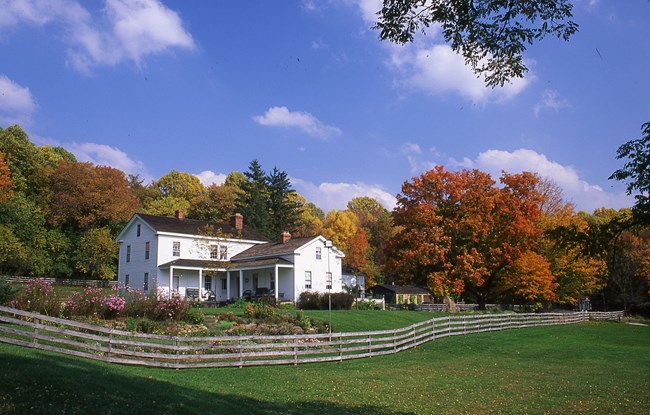 Exterior of a white, two-story building; trees with multi-colored leaves behind, and in front, a garden and wooden fence.