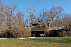 A brown wood building with brown shingle roof, surrounded by a green lawn and trees in the background.