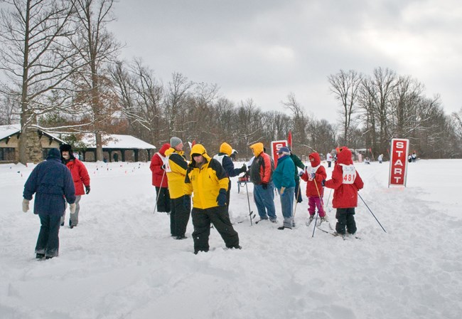 A group of cross country skiers and coaches stand, in full winter clothing, at the snow covered start line of a cross country ski race during the Special Olympics. A group of cross country skiers and coaches stand, in full winter clothing, at the snow covered start line of a cross country ski race during the Special Olympics.
