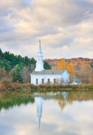 A white building with white steeple is reflected in a body of water; green and orange trees in the background.