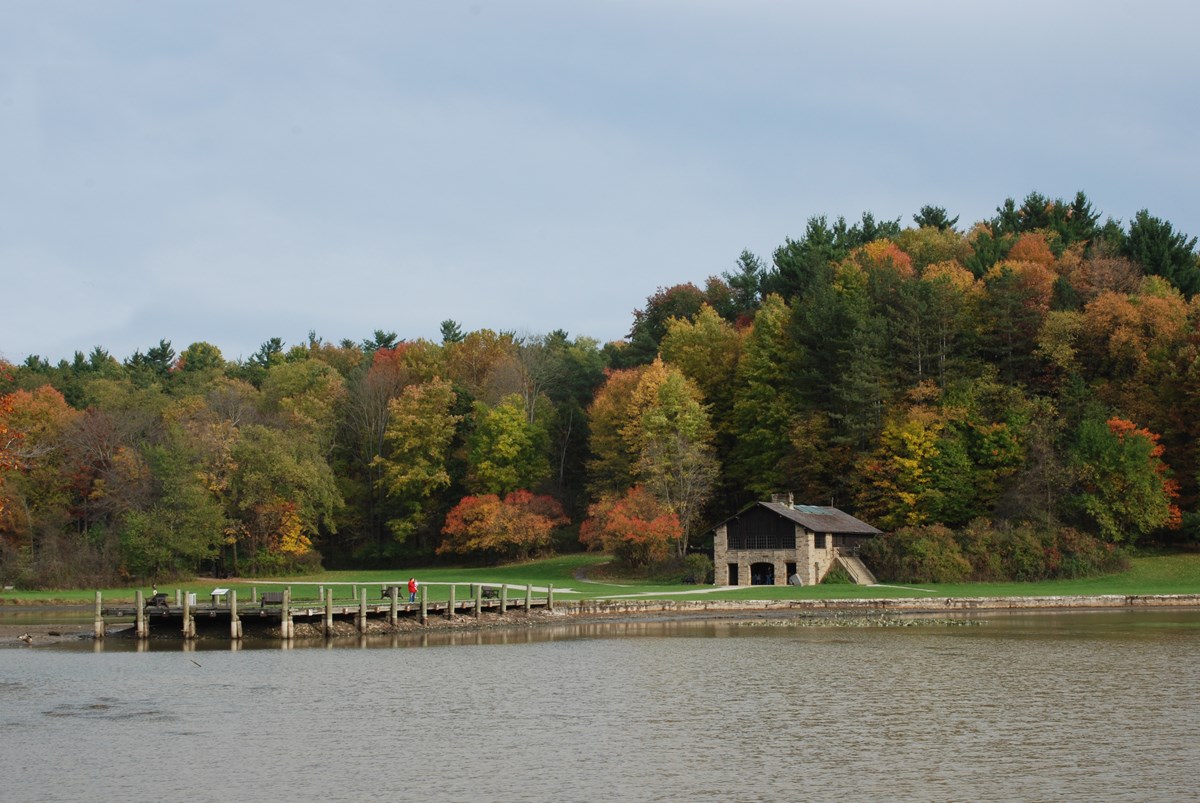 A rustic shelter made of stone and wood sits at the end of a long fishing pier that juts into a calm lake. Beyond the green grass by the building is a forested hillside made colorful by autumn leaves.
