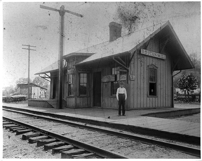 A man stands in front of a building labeled "Boston Mill"; train tracks in the foreground.
