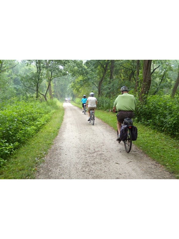 Three bikers ride down the towpath trail. The surrounding area is wooded and full of green foilage.