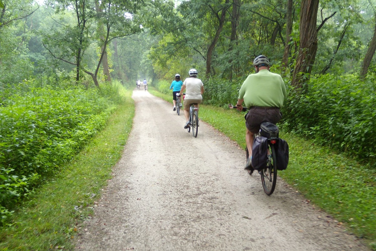 Three bikers ride down the towpath trail. The surrounding area is wooded and full of green foilage.