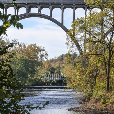 An arched concrete bridge crosses high over a river with trees along its banks; in the distance, a smaller white metal bridge crosses much closer to the water.