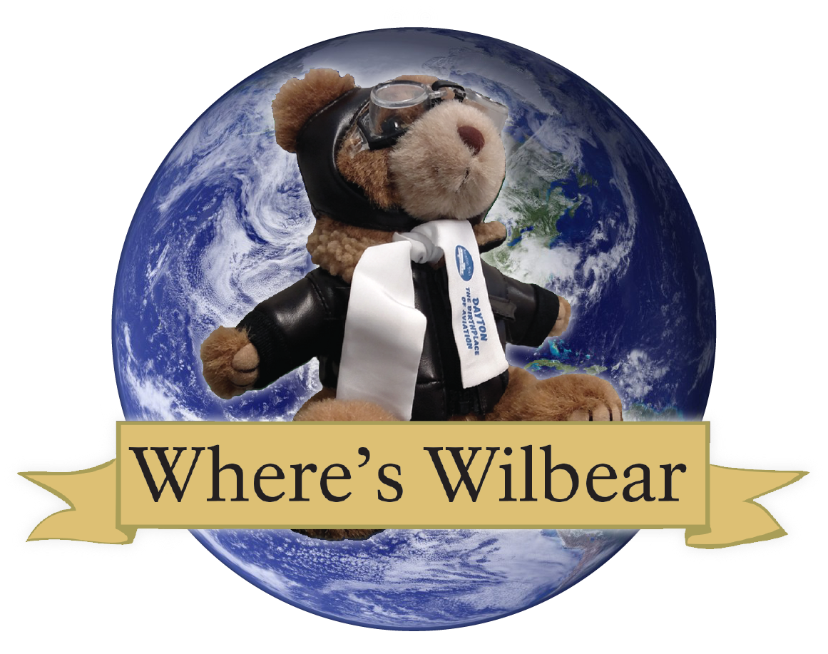 A stuffed bear wearing a brown jacket and eye goggles in front of an image of the world. A stuffed bear wearing a brown jacket and eye goggles in front of an image of the world.