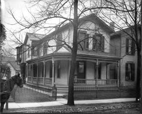 A two-story home with a large, wrap-around porch with a sidewalk and trees in the front yard.
