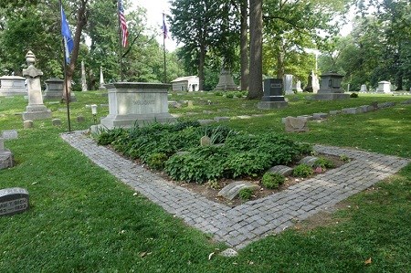 A burial plot surrounded by stone pavers and decorated with green vegetation and a large headstone.