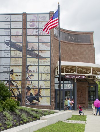 An American flag waves on top of a pole in front of a brick building as people enter