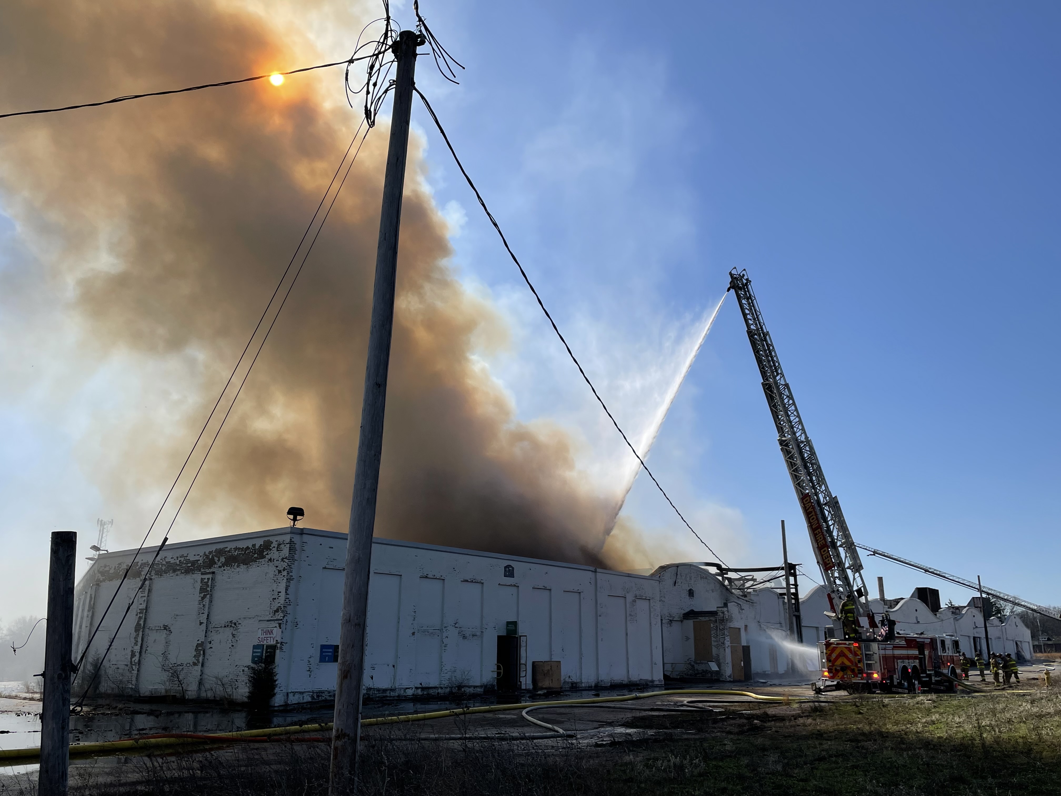 A white brick building is on fire with smoke rising and blocking the sun's rays as a fire crew works at a truck to launch water into the building from a ladder extended into the air.