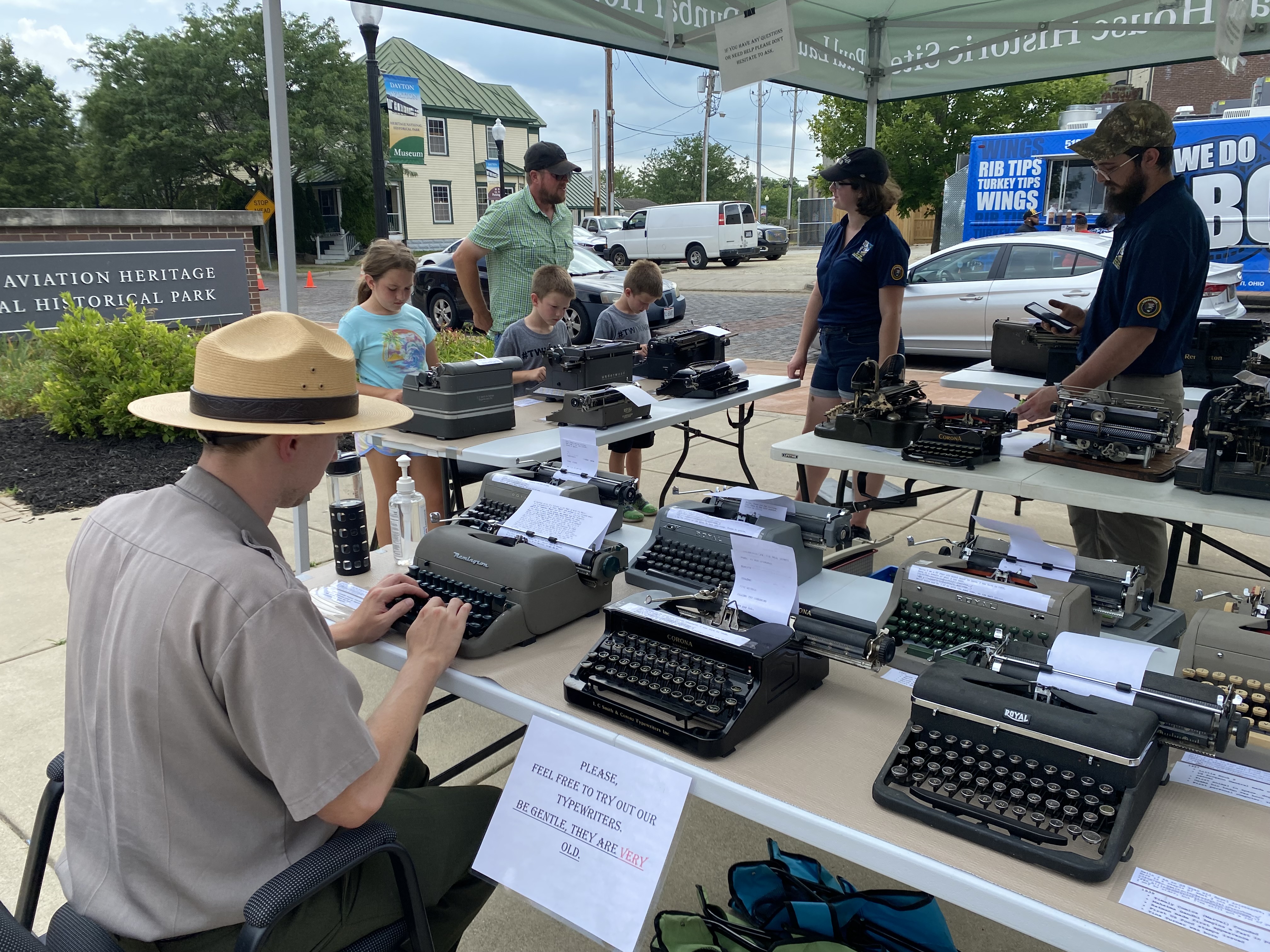 A ranger sits at a desk typing on a typewriter as volunteers talk with a family about park history.