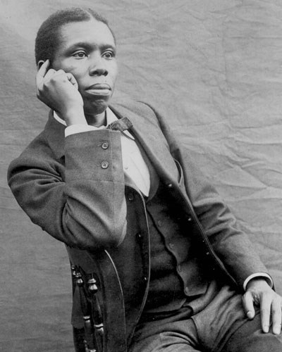 Paul Laurence Dunbar posing for a portrait with his hand rested against his face as he sits leaning his right side against a wooden chair.